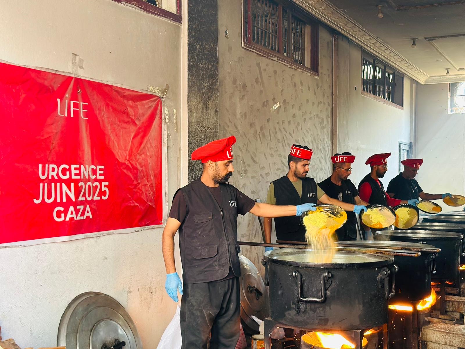Des membres de LIFE en casquettes rouges préparent un repas dans de grandes marmites, sous une affiche Urgence Gaza.